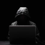 A hooded figure sitting in front of a laptop in a dark setting