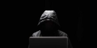 A hooded figure sitting in front of a laptop in a dark setting