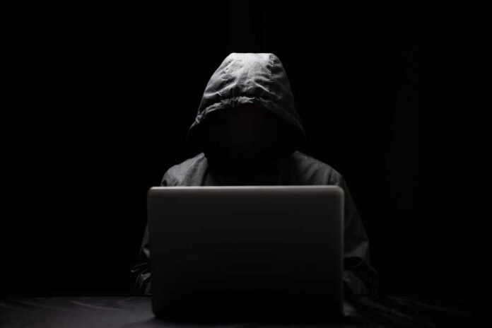 shutterstock_2072786747.jpg A hooded figure sitting in front of a laptop in a dark setting