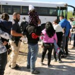 Families and individuals waiting in line near a bus with border patrol officers present