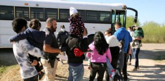 Families and individuals waiting in line near a bus with border patrol officers present