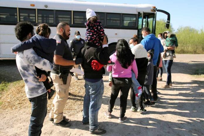 shutterstock_2091429184.jpg Families and individuals waiting in line near a bus with border patrol officers present