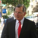 Barrasso Blasts Democrats: Illegal Immigrant Perks? A man in a suit with a red tie walking outdoors, accompanied by a police officer