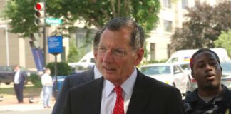 A man in a suit with a red tie walking outdoors, accompanied by a police officer