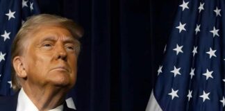 A political figure looking confidently while standing near American flags