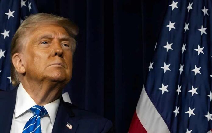 A political figure looking confidently while standing near American flags
