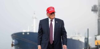A man in a suit wearing a red cap stands near a ship at a harbor