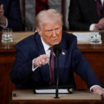 Trump’s “Hall Of Shame” Targets Influencers Man in suit speaking at a podium during an event