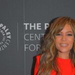 Woman with wavy hair in a bright red dress at media event backdrop